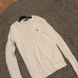 Vit stickad tröja från Ralph Lauren - Säljer en klassisk vit stickad tröja från Ralph Lauren med kabelstickat mönster. Tröjan har långa ärmar och en liten broderad logotyp på bröstet. Perfekt för kyligare dagar! Nypris 1900kr. Ska tvättas innan leverans! 