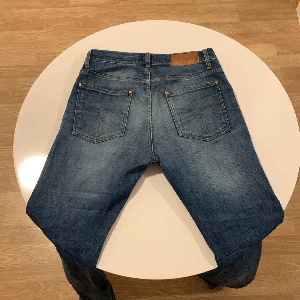 Snygga blå jeans från Tiger of Sweden med klassisk femficksdesign och slitna detaljer. De har en knappgylf och är tillverkade i ett robust denimtyg. Perfekta för en avslappnad stil.. Farkut & Housut.