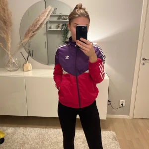 Färgglad vindjacka från Adidas - Säljer en cool vindjacka från Adidas i lila och röd med vita detaljer. Jackan har dragkedja och de klassiska tre ränderna längs ärmarna. Perfekt för en sportig look eller en avslappnad dag ute. 🌀