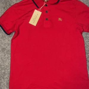 Röd pikétröja från Burberry - Snygg röd pikétröja från Burberry med klassisk krage och knappar framtill. Tröjan har en liten broderad logga på bröstet och kontrasterande detaljer vid kragen. Perfekt för en stilren look.