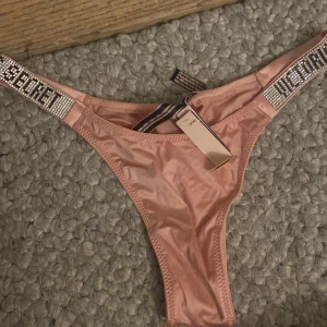 Rosa trosor från Victoria's Secret - Snygg rosa bikiniunderdel från Victoria's Secret med glittriga detaljer och text på banden. Den har en modern och stilren design som verkligen sticker ut. Helt ny 