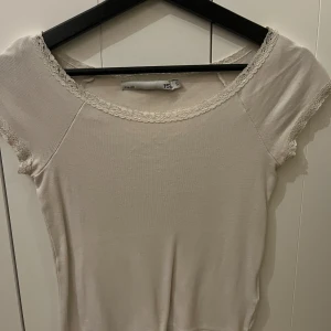 Vit topp med spetsdetaljer - Säljer en söt vit topp med spetsdetaljer runt halsringningen och ärmsluten. Toppen har korta ärmar och en mjuk, bekväm passform. Perfekt för en avslappnad stil. Använd ett par gånger, de ända är att den vita färgen på tröjan har blivit lite gul aktig i tonen