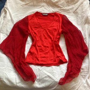 Röd blus med vida ärmar  - Säljer en snygg röd blus med vida, transparenta ärmar och v-ringning. Perfekt för att sticka ut med sin unika design och livfulla färg.