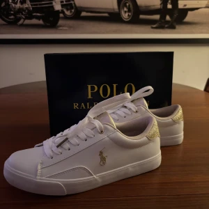 Vita sneakers från Polo Ralph Lauren - Säljer dessa helt orörda snygga vita sneakers från Polo Ralph Lauren med gulddetaljer. Skorna har snörning och en stilren design med märkets ikoniska logga på sidan. Perfekta för en trendig look.