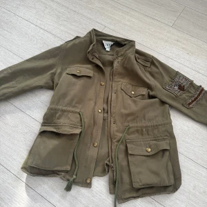 Olivgrön jacka från CiminY Paris - Säljer en olivgrön jacka från CiminY Paris med coola broderade detaljer på ärmen. Jackan har flera fickor med knappar och en dragkedja framtill. Perfekt för en avslappnad stil.