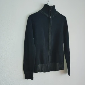 Svart kofta från Identity - Säljer en stilren svart kofta från Identity med dragkedja framtill. Koftan har en ribbad krage och ribbade muddar vid ärmslut och nederkant, vilket ger en snygg och bekväm passform. Perfekt för kyligare dagar.