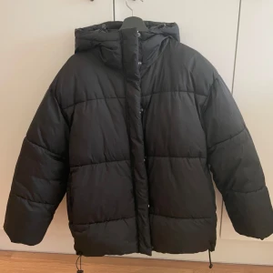 Svart pufferjacka från Monki - Säljer en svart pufferjacka från Monki med huva och dragkedja. Jackan har en oversized passform och är perfekt för kalla dagar. Den är stilren och enkel att matcha med olika outfits.