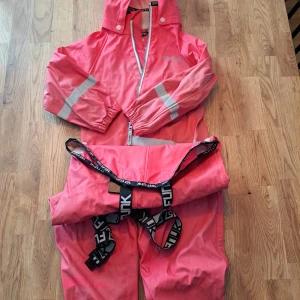 regnjacka från Funk - Säljer en rosa regnjacka från Funk i storlek 110. Jackan har en huva och dragkedja framtill samt reflexdetaljer på ärmarna. Materialet är 100% polyester vilket gör den perfekt för regniga dagar. Hängslen ingår för extra komfort och passform.