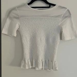 Vit topp från Gina Tricot - Säljer en söt vit topp från Gina Tricot med korta ärmar och volangdetalj vid nederkanten. Toppen har en smockad design framtill som ger en fin passform. I storlek 146/152 och sitter som xs/s.