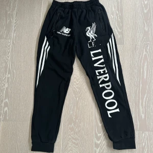 Svarta träningsbyxor från New Balance - Snygga svarta träningsbyxor med Liverpool FC-logga och New Balance-märke. Byxorna har elastisk midja och dragkedjefickor. Perfekta för fotbollsfans! Det är bara en defekt och det är att "C" har lossnat till L.F.C. men det är inget man tänker så mycket på. Vet inte vad det är för storlek då lappen är avklippt men uppskattar till S. Nypris är 1200kr