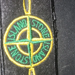 Svart Loga från Stone Island - Snygg logo jacka från Stone Island med det ikoniska kompassmärket i grönt och gult. Perfekt för dig som vill ha en stilren och trendig look.