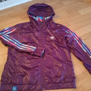 Lila vindjacka från Adidas - Säljer en cool lila vindjacka från Adidas med huva och dragkedja. Jackan har ett färgglatt geometriskt mönster på insidan och längs ärmarna. Perfekt för att sticka ut i mängden! 💜