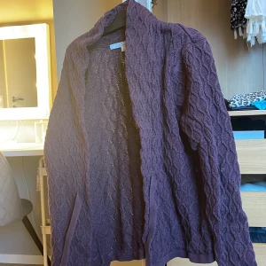 Lila stickad kofta från First Class - Säljer en mysig lila stickad kofta från First Class. Koftan har ett fint kabelstickat mönster och långa ärmar. Perfekt för kyliga dagar. Den har även ett knytband i midjan för en justerbar passform. Passar mig som är i storlek xs/s