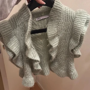 Grön bolero zara - Säljer en mysig grön stickad kofta från Zara med volangdetaljer. Koftan har en unik design med korta ärmar och ribbade kanter. Perfekt för att hålla sig varm och stilren under kyligare dagar.