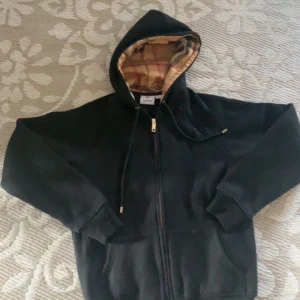 Svart hoodie från Burberry - Säljer en stilren svart hoodie från Burberry med dragkedja och en klassisk rutig mönstrad huva. Perfekt för en avslappnad och trendig look. Storlek M men passar XS. Pris kan diskuteras 