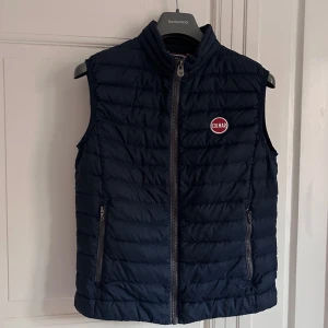 Mörkblå dunväst från Colmar - Snygg mörkblå dunväst från Colmar med dragkedja framtill och två praktiska fickor med dragkedjor. Västen har en quiltad design och en rund logotyp på bröstet. Perfekt för kyligare dagar. Skicket är mycket bra och hör er vid frågor.