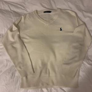 Vit kashmirtröja från Ralph Lauren - Säljer en elegant vit kashmirtröja från Ralph Lauren med v-ringning och broderad logotyp på bröstet. Tröjan har långa ärmar och ribbade muddar vid ärmslut och nederkant. Perfekt för en stilren look.