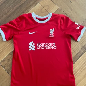 Äkta Liverpool fotbollströja- Från säsongen 2023-2024 - Säljer en röd Liverpool fotbollströja från Nike med vit logga och sponsortryck. Tröjan har korta ärmar med vita detaljer och en rund halsringning. Perfekt för alla Liverpool-fans! 🔴
