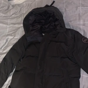 Svart dunjacka från Canada Goose - Säljer en svart dunjacka från Canada Goose med huva och broderad logga på ärmen. Jackan har en stilren design med långa ärmar och är perfekt för kalla dagar. Den är tillverkad i slitstarkt material och har en bekväm passform. Perfekt för vintern!  Passar perfekt med längden 180cm