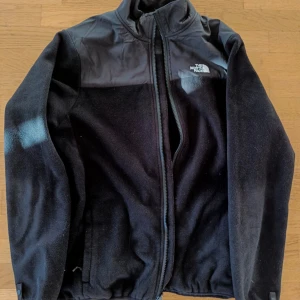 Svart North Face fleece - Säljer en North Face fleece i storlek L med dragkedja och hög krage. Jackan har en broderad logga på bröstet och axeln. Sparsamt använd och bra kvalitet. Säljes då den är lite kort för mig. Själv är jag 191 cm, 74 kg, har ofta M-L på hoodies men denna passar snarare som S-M. Kan ej verifiera dess äkthet och säljes i befintligt skick.