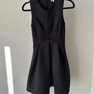 Svart klänning från Elly Pistol - Snygg svart klänning från Elly Pistol i en stilren design. Klänningen är ärmlös och har en figurnära passform med en subtil skärning i midjan. Perfekt för en elegant look.