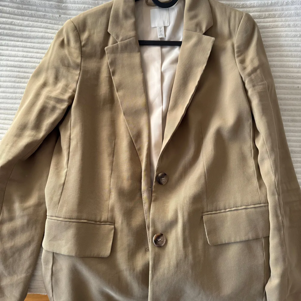 Snygg beige kavaj från H&M med klassisk design och två knappar framtill. Kavajen har långa ärmar och två fickor med lock. Perfekt för en stilren look.. Takit.