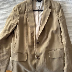 Beige kavaj från H&M - Snygg beige kavaj från H&M med klassisk design och två knappar framtill. Kavajen har långa ärmar och två fickor med lock. Perfekt för en stilren look.
