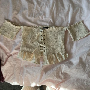 Beige offshoulder topp från Zara - Säljer en söt beige offshoulder topp från Zara med snörning framtill och volangdetaljer vid nederkanten. Toppen har en smockad design som ger en fin passform. Perfekt för en somrig look! Endast använd vid ett tillfälle så den är som ny. Skriv om ni har några frågor! 💞