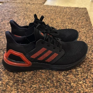 Svarta och röda Adidas sneakers - Snygga Adidas sneakers i svart med röda detaljer. Skorna har en stickad ovandel och klassiska tre ränder på sidan. De är utrustade med snörning och har en bekväm sula för extra komfort. Nypris 1800kr