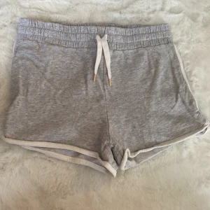 Grå shorts från H&M - Säljer ett par grå mjuka shorts från H&M med vit snörning i midjan. Perfekta för en avslappnad dag hemma eller lätt träning. De har en elastisk midja och vita kantdetaljer som ger en sportig look.