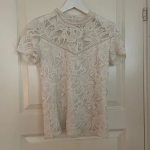 Vit spetstopp - Elegant vit spetsblus med korta ärmar och ett vackert blommönster. Blusen har en rund hals och är perfekt för en stilren look. Den genombrutna spetsen ger en luftig och feminin känsla.