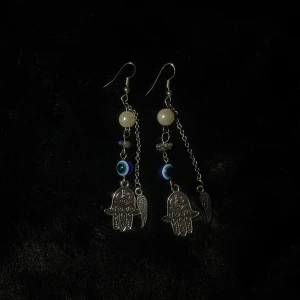 Silverfärgade örhängen med hamsa och pärlor - Handgjorda unika silverfärgade örhängen med hamsa-hängen och dekorativa pärlor. Designen inkluderar även blå och vita detaljer samt små fjäderhängen. Perfekta för att ge en vibe touch till din stil. 