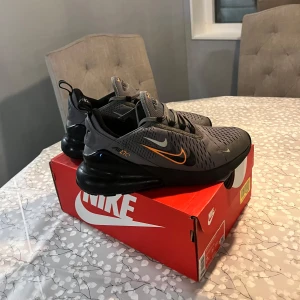  Nike Air Max 270 - Snygga grå Nike Air Max 270 sneakers med orange detaljer. Skorna har en modern design med synlig Air-enhet i hälen för extra komfort. Perfekta för både stil och funktion.