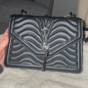 Svart  axelväska från YSL - Snygg svart axelväska från YSL med quiltat mönster och silverdetaljer. Väskan har en elegant kedjerem och en dekorativ tofs framtill. Knappt använd och säljer pga att den ej kommit till användning 🌸