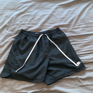 Svarta badshorts från Nike - Snygga svarta shorts från Nike med vit snörning i midjan. Perfekta för en dag vid poolen eller en avslappnad dag på stranden. De har en enkel design med en liten Nike-logga på benet.