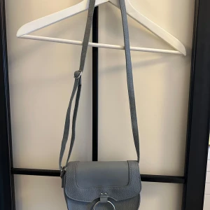 Grå axelväska från Primark - Snygg grå axelväska från Primark med justerbar axelrem och stilren design. Perfekt för att bära dina dagliga nödvändigheter med stil. Väskan har en praktisk stängning och en modern look.