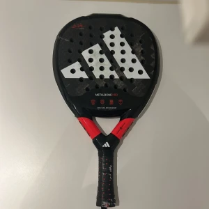 Padelracket Adidas Metalbone HRD - Säljer ett Adidas Metalbone HRD padelracket. Racketen har en svart och röd design med vita detaljer och är utrustad med strukturell förstärkning för bättre hållbarhet. Perfekt för den som söker hög prestanda på padelbanan. Har sprickor i racket som förändrar spelet rätt så mycket. Är absolut villig och negotiera.