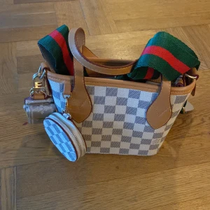 Rutig handväska med axelrem - Snygg handväska med rutigt mönster i beige och vitt. Väskan har bruna skinnhandtag och en avtagbar axelrem i grönt och rött. Perfekt för att ge en färgklick till din outfit.