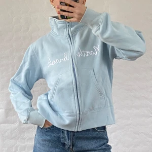 Ljusblå fleecejacka med broderi - Mysig ljusblå fleecejacka med dragkedja och broderad text på bröstet. Jackan har en hög krage och långa ärmar, perfekt för kyliga dagar. Praktiska fickor framtill för extra komfort.