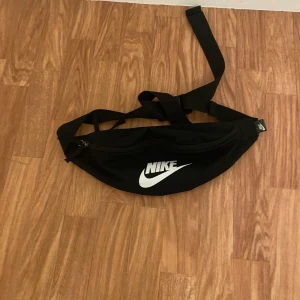 Svart midjeväska från Nike - Säljer en svart midjeväska från Nike med justerbart band och dragkedja. Perfekt för att hålla dina saker säkra och nära till hands. Den har ett stort fack med nätficka inuti för extra förvaring.