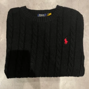 Svart kabelstickad tröja från Polo Ralph Lauren - Snygg svart kabelstickad tröja från Polo Ralph Lauren med det ikoniska röda logotypbroderiet på bröstet. Tröjan har en klassisk rund halsringning och långa ärmar. Perfekt för en stilren look.