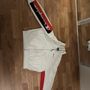 Vit jacka från Champion - Säljer en vit jacka från Champion med dragkedja och ribbade muddar. Jackan har röda och svarta detaljer på ärmarna och Champion-loggan broderad på bröstet. Perfekt för en sportig look!