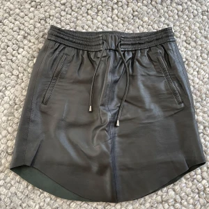 Svart skinnkjol från Second Female - Snygg skinnkjol från Second Female med elastisk midja och dragsko. Tillverkade i 100% läder med polyesterfoder för extra komfort. Fickor i sidorna. 