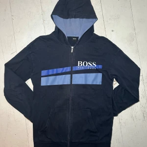 Svart hoodie från Hugo Boss - Snygg svart hoodie från Hugo Boss med dragkedja och blå detaljer på bröstet. Hoodien är i storlek M och är i bra skick. Tveka inte på att höra av dig ifall du har några som helst frågor, jag svarar snabbt 💪👑