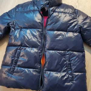 Mörkblå pufferjacka från Tommy Hilfiger - Snygg mörkblå pufferjacka från Tommy Hilfiger med dragkedja och hög krage. Jackan har märkets logga på bröstet och stor text på ryggen. Perfekt för kyliga dagar med sin vadderade design.