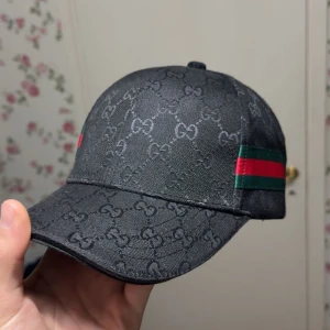 Gucci keps - Snygg svart keps från Gucci med det ikoniska GG-mönstret. Kepsen har gröna och röda ränder på sidan, vilket ger en klassisk touch. Perfekt för att lyfta vilken outfit som helst.