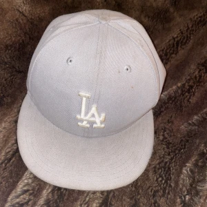 Grå keps från New Era - Snygg grå keps från New Era med LA-logga framtill. Perfekt för att komplettera din streetwear-look. Kepsen har en klassisk design med platt skärm och med en vit broderad logotyp.
