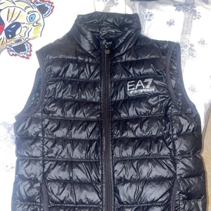 Svart dunväst från Emporio Armani - Snygg svart dunväst från Emporio Armani med EA7-logga på bröstet. Västen har en praktisk dragkedja framtill och är perfekt för kyligare dagar. Den quiltade designen ger en stilren look.