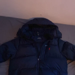Mörkblå dunjacka från Polo Ralph Lauren - Säljer en snygg mörkblå dunjacka från Polo Ralph Lauren med huva och dragkedja. Jackan har en liten röd logotyp på bröstet och är perfekt för kyliga dagar.