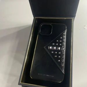 Svart mobilskal från iDeal of Sweden - Snyggt svart mobilskal från iDeal of Sweden med stilrena nitar i silver. Skalet har en unik design med en triangulär detalj som ger det en modern touch. Perfekt för att ge din mobil en trendig look. Säljs på grund av fel storlek till min mobil ALDRIG ANVÄND 